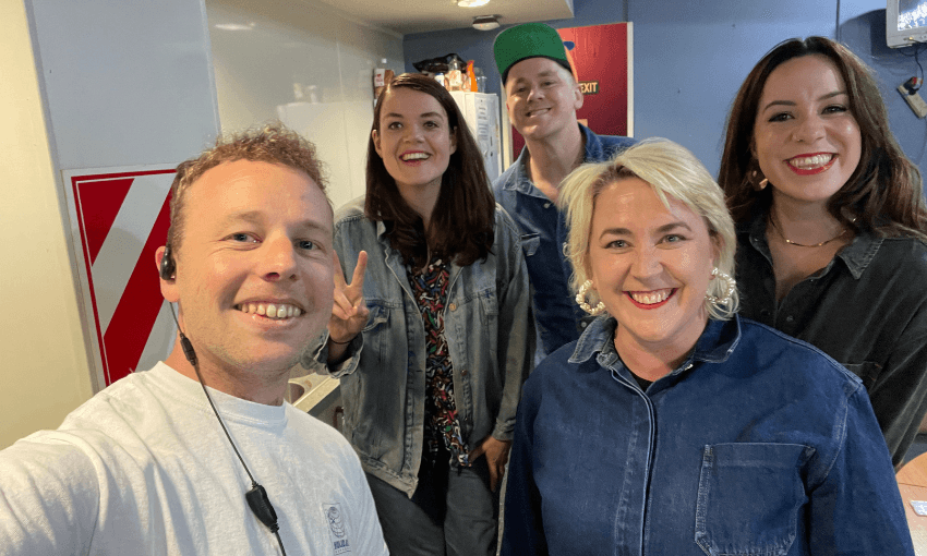Five smiling people pose for a selfie indoors. One person holds up a peace sign. They are casually dressed, most in denim, and appear to be in a friendly, relaxed setting.
