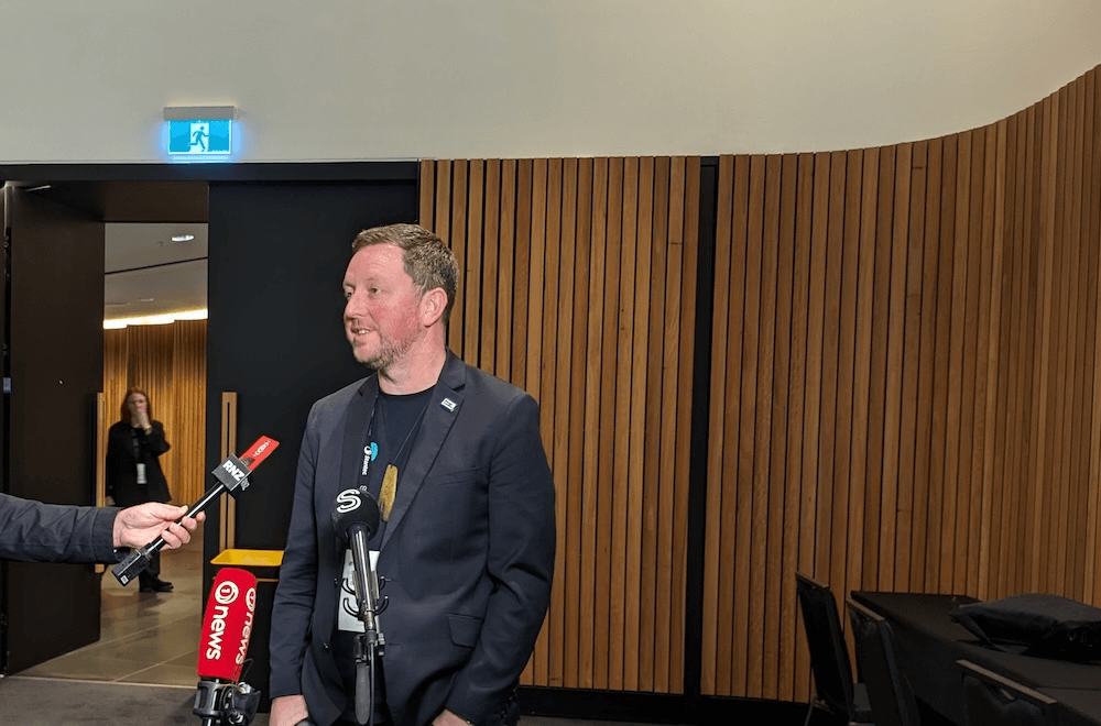 Sam broughton, a cheerful guy in a blazer, talks with wood panelling behind him and lots of microphones in front 