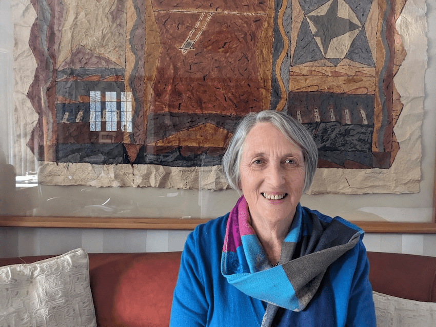 Kate Dewes, an older woman with grey hair and a colourful scarf, sits on a red/brown sofa with some earth-toned art behind her and a slight smile