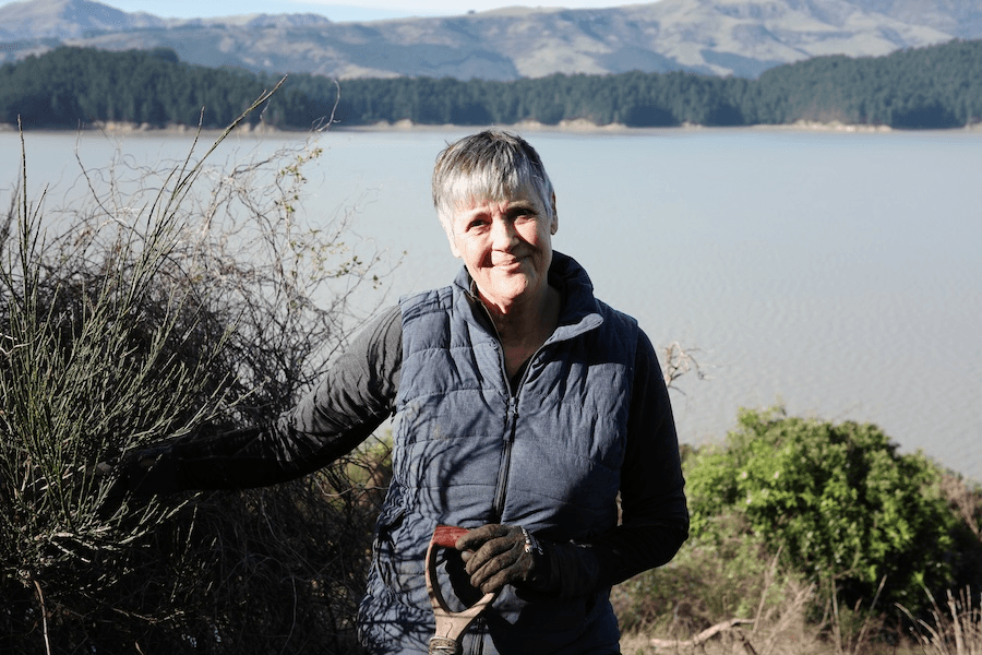 a grey haird woman wearing a navy vest and holding a spade with the sea behind her