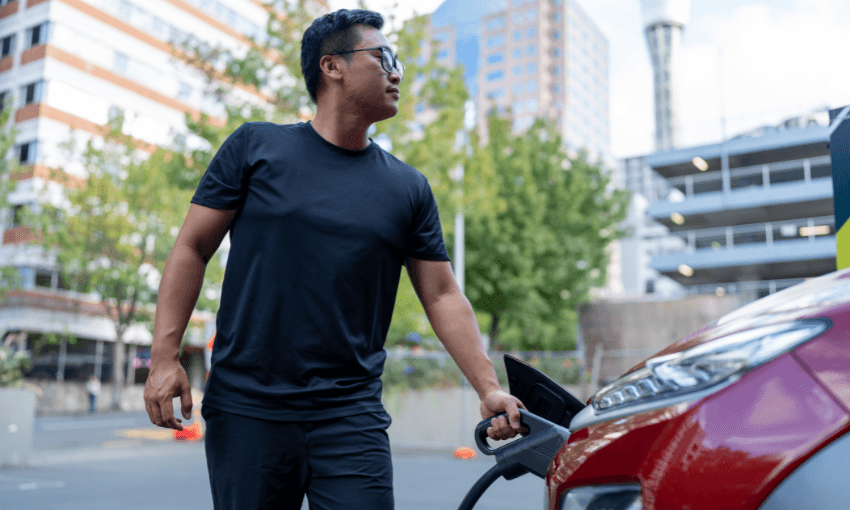 A man wearing glasses and a black shirt is charging a red electric car at an outdoor charging station in an urban area with buildings and trees in the background.