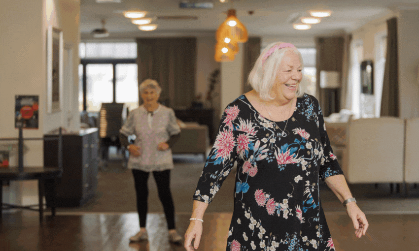 Two older women are smiling and walking inside a brightly lit room; one woman in a black floral dress is in the foreground, while another in casual clothes stands further back.