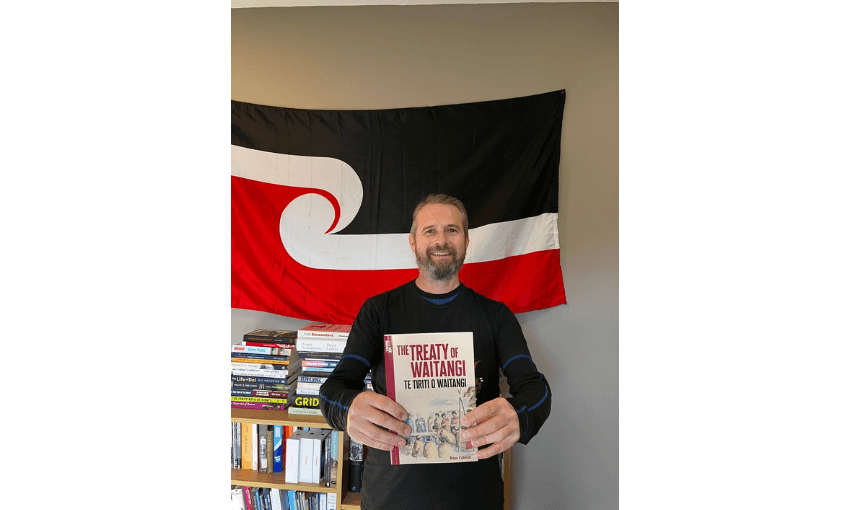 A photograph of a man holding up a book titled The Treaty of Waitangi with a tino rangatiratanga flag behind him and bookshelves.o