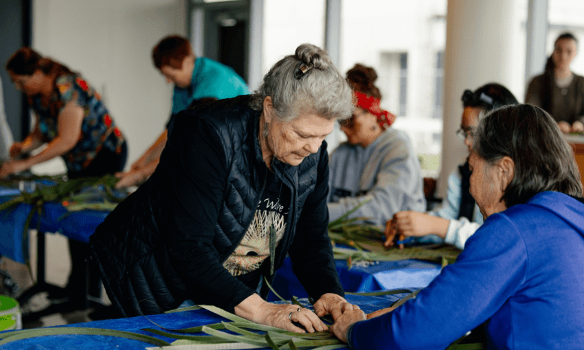 a group of people bending over a table weaving green harakeke in natural light