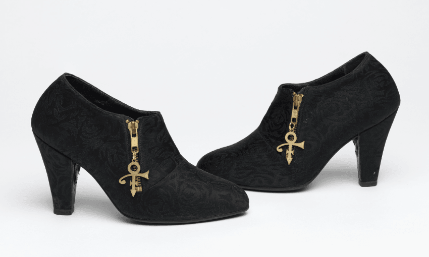 A pair of black heeled ankle boots with subtle floral patterns and gold zippers featuring the Prince "Love Symbol" as zipper pulls, displayed on a white background. Prince's shoes.