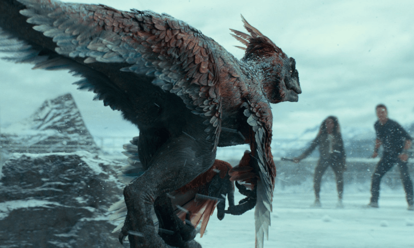 Still of a part of a Jurassic Park movie where a huge bird like dinosaur is chasing two people in the distance. It is icy and snowy terrain. 