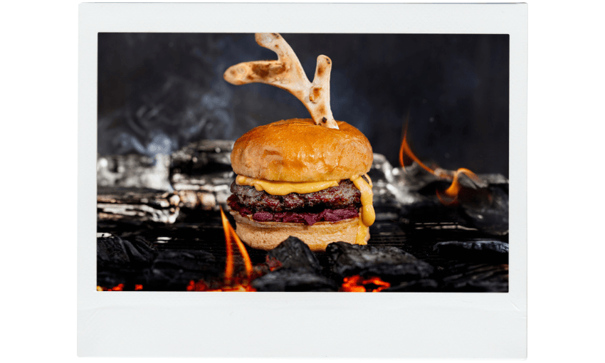 A cheeseburger with melted cheese sits on a grill above glowing coals, topped with two bread pieces shaped like antlers. Flames and smoke rise in the background, creating a dramatic effect.