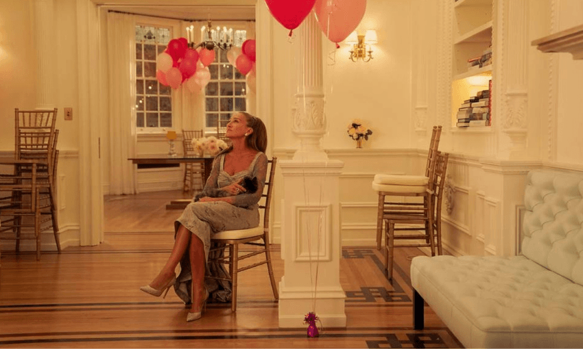 A woman in a silver dress sits alone on a chair in an elegant, warmly lit room decorated with pink and red balloons. She gazes upward, surrounded by empty chairs and a white couch, with a table of balloons in the background.