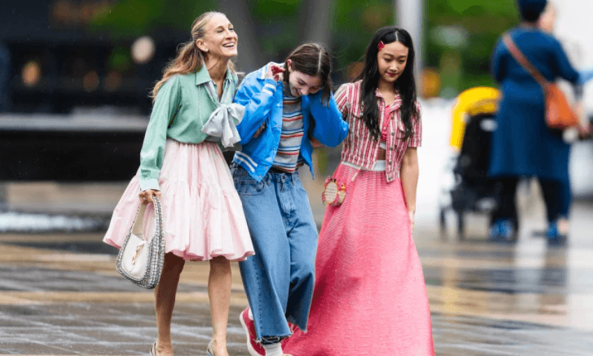 Three women walk together outdoors, smiling and laughing. They wear colorful, stylish outfits, including skirts and jeans, and appear to be enjoying each other's company on a city street.