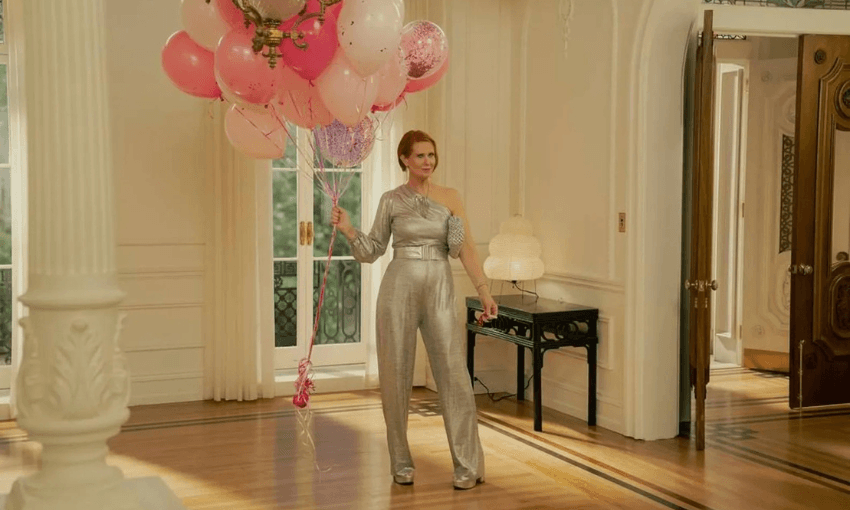 A woman in a shiny silver jumpsuit stands in a bright, elegant room holding a large bunch of pink and white balloons. Sunlight streams through tall windows, and the room features classic white decor and wooden floors.