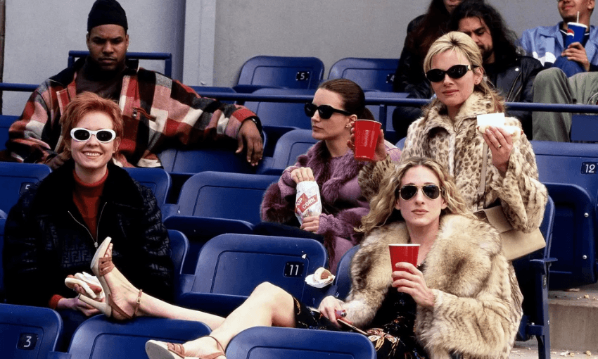 Four stylish women sit in stadium seats, wearing sunglasses and fashionable coats, holding drinks and snacks, with empty blue seats around them and other people in the background.