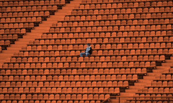 A lone person sits among many empty orange seats in a large stadium, appearing small and isolated in the vast, deserted space.