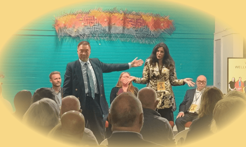 Two presenters stand in front of a seated audience, gesturing expressively. Three other panelists are seated behind them. The background features a teal wall and colorful artwork. The scene has a soft yellow vignette effect.