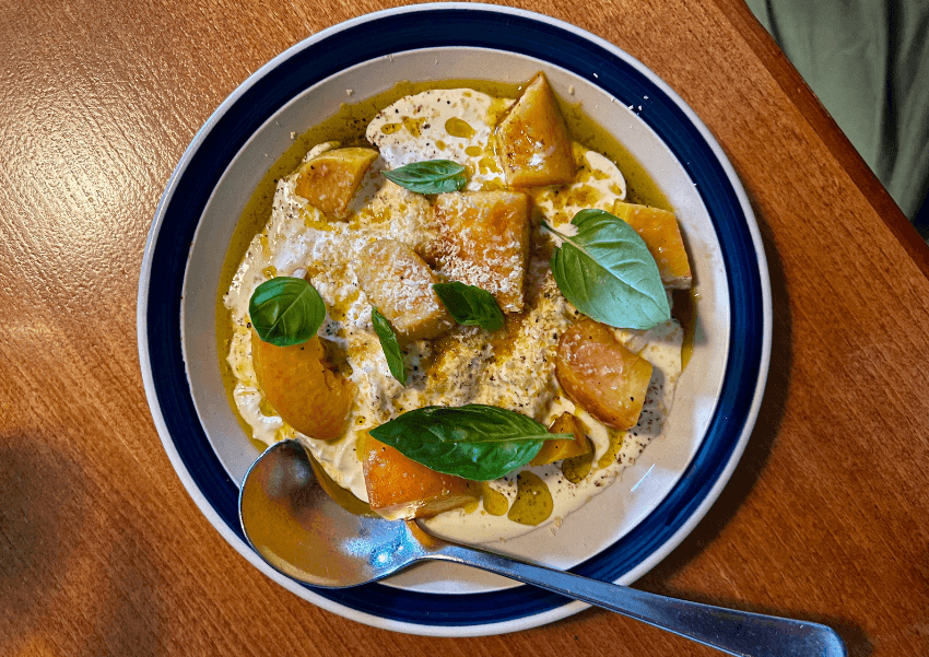 A bowl of creamy soup topped with golden roasted root vegetables, fresh basil leaves, grated cheese, and a drizzle of oil sits on a wooden table with a metal spoon resting on the side of the dish.