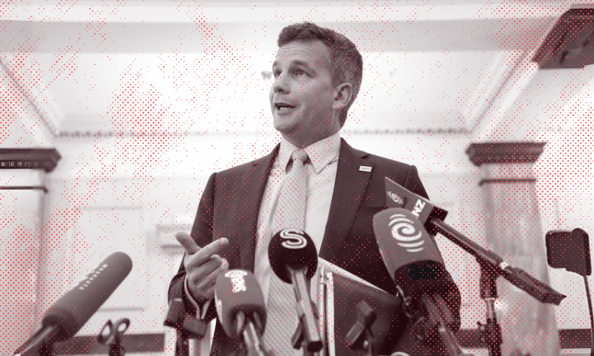 A man in a suit speaks at a press conference, standing behind several microphones from various news outlets in a formal indoor setting.