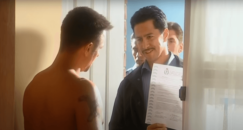 A shirtless man opens the door to a police officer holding an arrest warrant