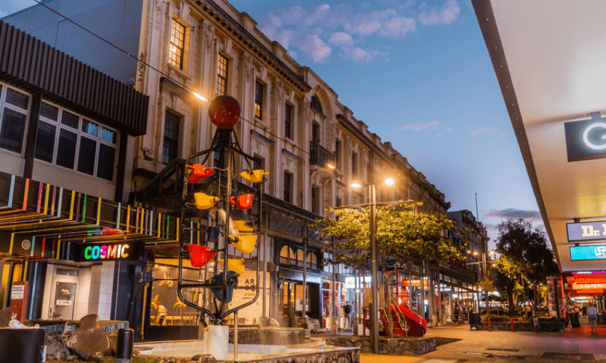 A lively city street at dusk with colorful shops, a whimsical bucket fountain in the center, illuminated buildings, and a partly cloudy sky overhead. People are visible strolling along the sidewalks.