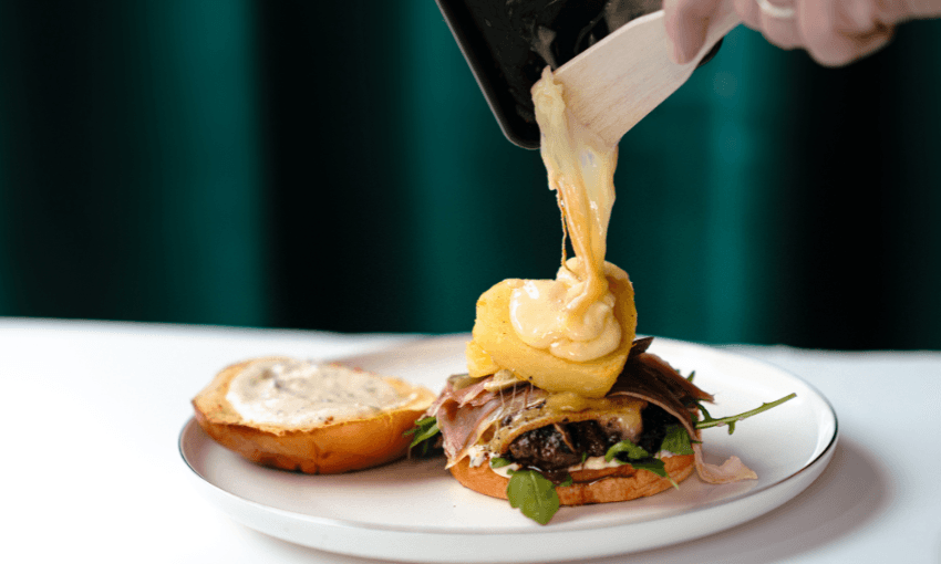 A hand uses a wooden spatula to pour melted cheese over a burger with greens and prosciutto on a white plate, with the top bun and sauce set aside.