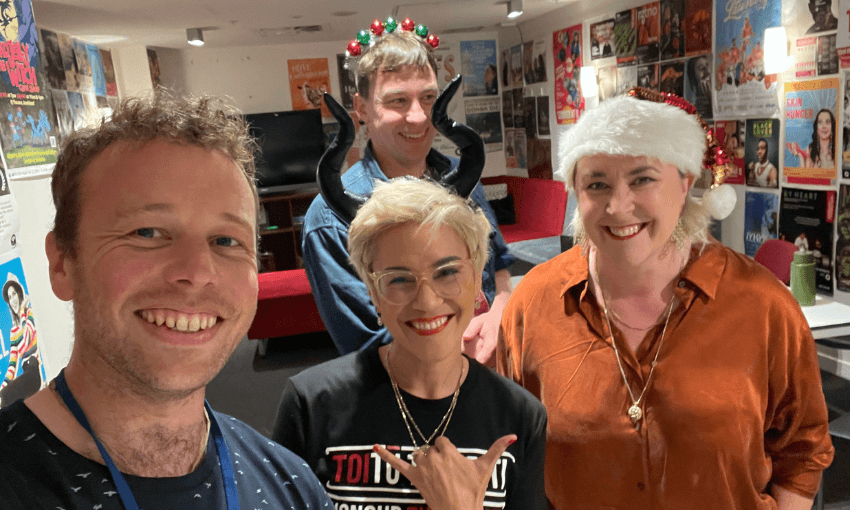 Four people smiling indoors, some wearing festive accessories: one has reindeer antlers, another black horns, and another a Santa hat. They stand in front of a wall covered with colorful posters.