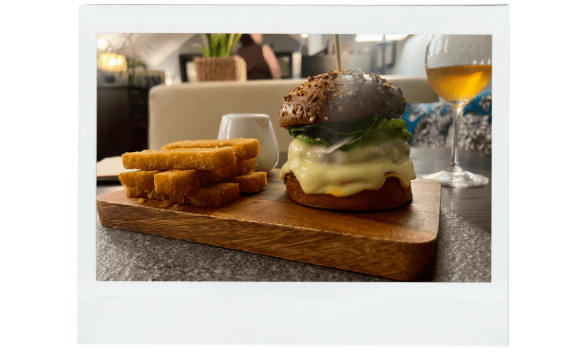 A wooden board with a cheeseburger topped with lettuce and a sesame bun, next to several thick, golden fries. A glass of drink sits in the background on a restaurant table.