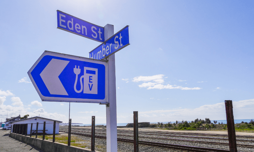 A blue road sign shows a left arrow with an electric vehicle charging symbol below street signs for Eden St and Humber St, near train tracks and a small building under a bright sky.
