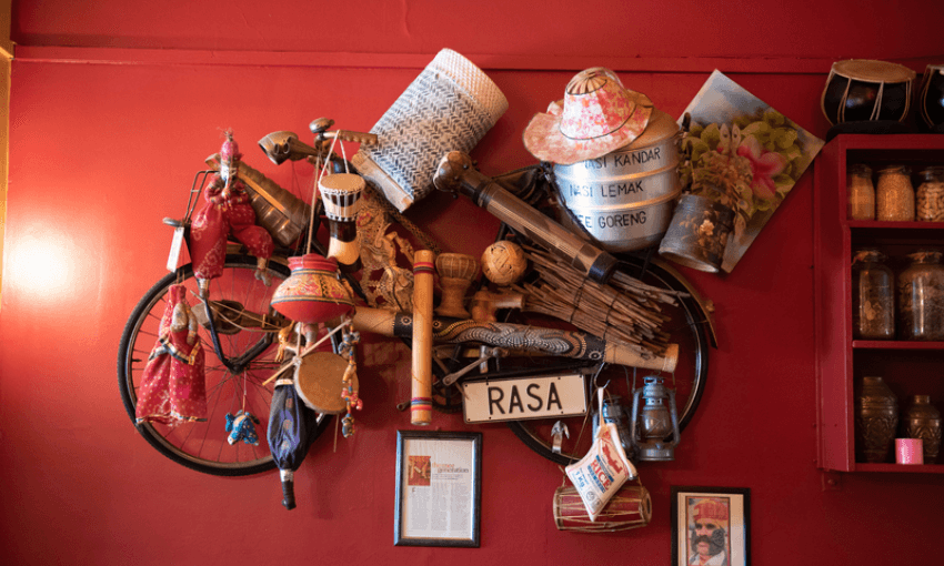 A red wall decorated with a vintage bicycle adorned with baskets, hats, musical instruments, a sign reading "RASA," framed photos, and jars filled with spices.
