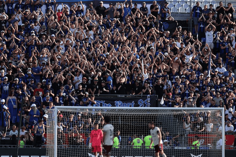 A large crowd of enthusiastic soccer fans in blue shirts cheer and raise their arms in unison in the stadium stands, while players and a goalkeeper are visible near the goal on the field.
