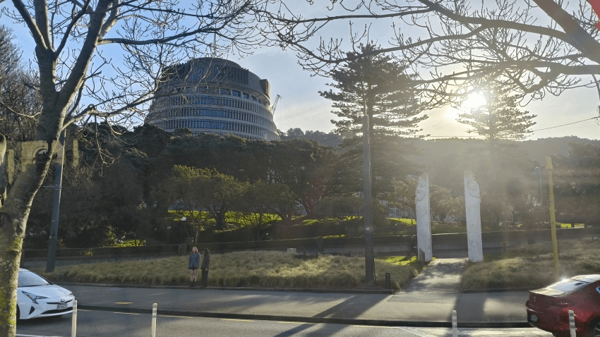 A round, modern government building sits atop a hill surrounded by trees, with sunlight streaming through. In the foreground, two people stand near a grassy area by a road, and three tall white sculptures are visible.