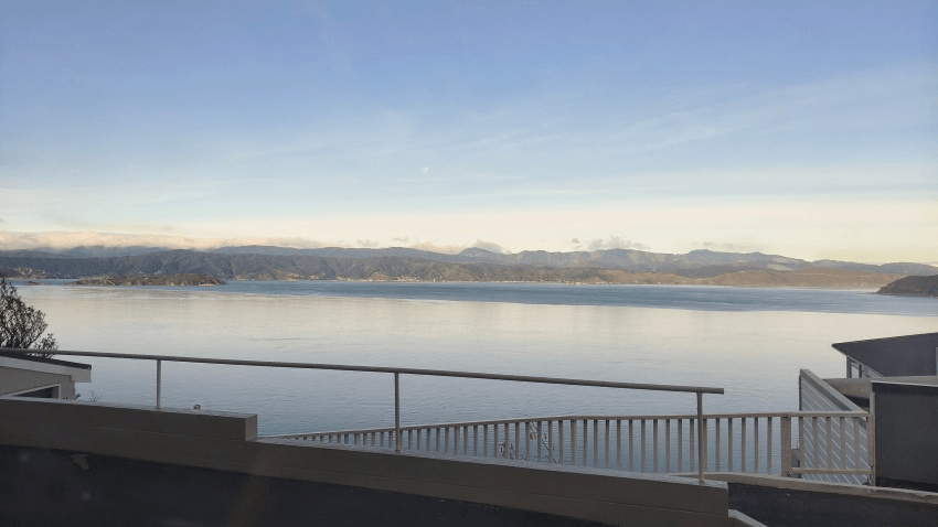 View of a calm bay with mountains in the distance, seen from behind a white railing on a balcony or terrace under a clear sky.