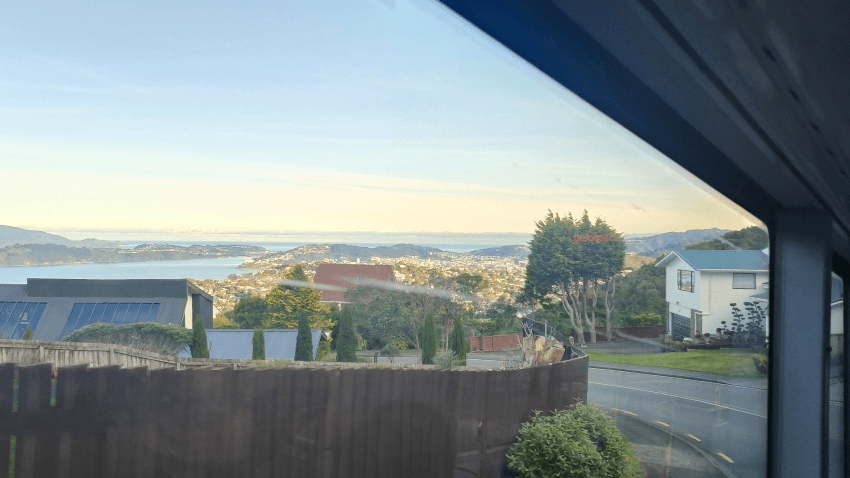 A scenic suburban neighborhood with houses, trees, and a winding road, overlooking a distant city and calm blue water under a clear sky, viewed through a window.