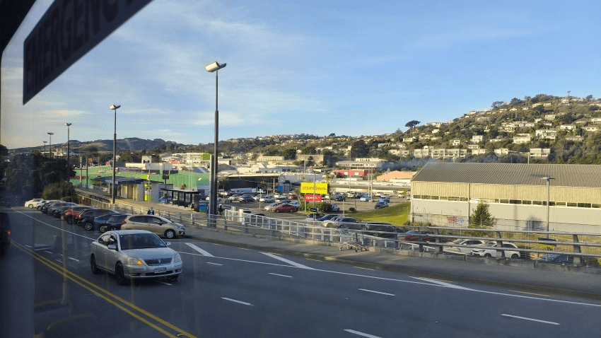 View from a bus window of a road with moving cars, a parking lot, and various buildings. In the background, there are hills with houses and clear skies above.