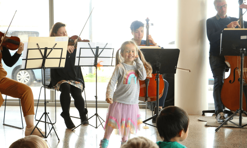 a girl in a tutu and a rainbow top dances in front of a cellist and violinist