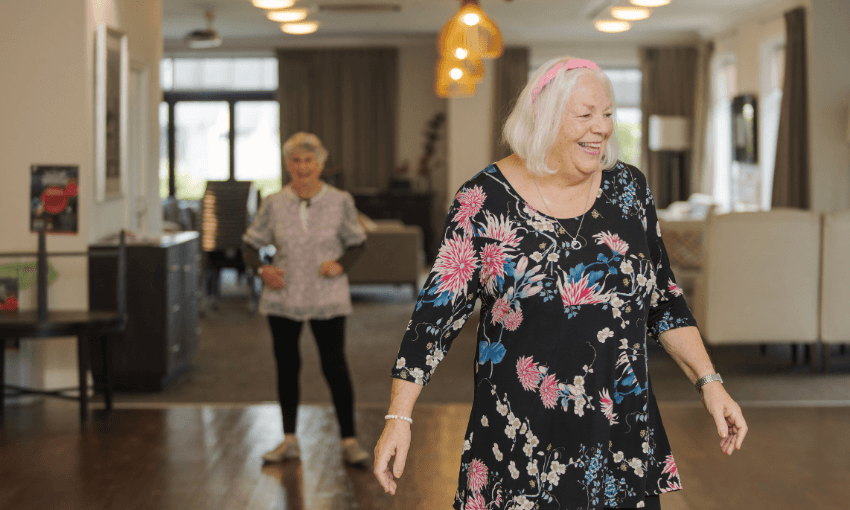 Two elderly women smile and dance in a bright, spacious living room. One woman in a black floral dress stands in the foreground, while the other is in the background, both enjoying the activity.