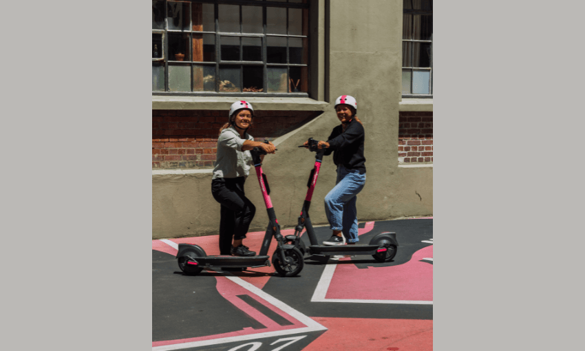two young women on pink-branded escooters in the sunshine, wearing helmets