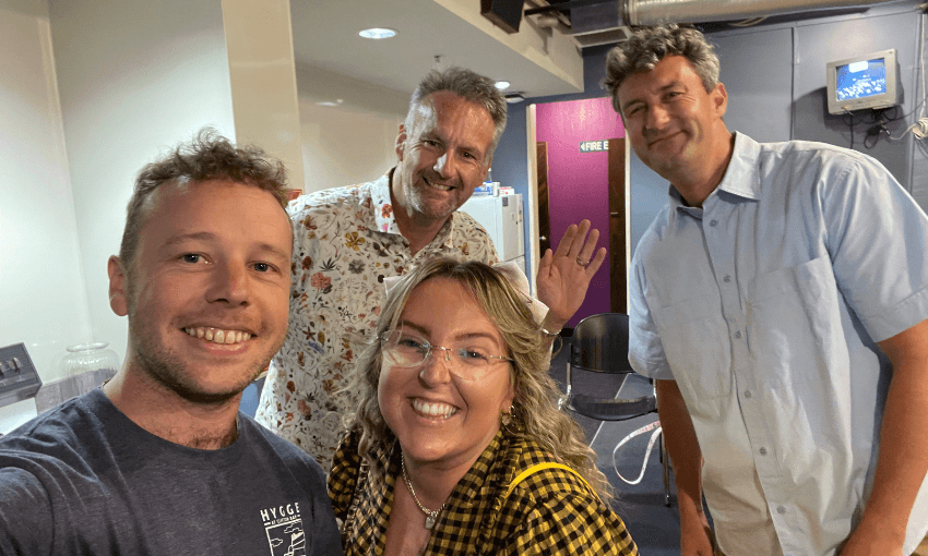 Four adults stand together indoors, smiling at the camera. Three men and one woman are in casual attire, with the woman wearing glasses and a yellow plaid shirt. The background shows a purple door and some office furniture.