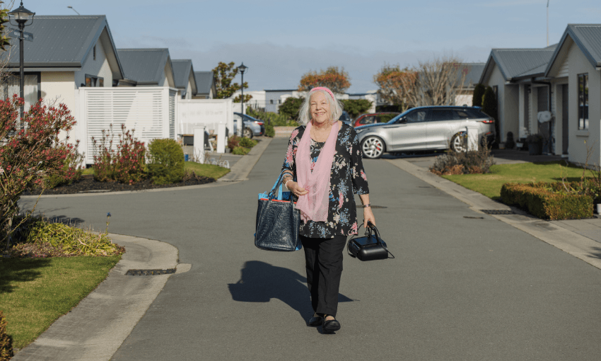 An older woman with white hair, dressed in black and pink, walks down a suburban street carrying shopping bags. Houses, gardens, and parked cars line the sunny neighborhood road.