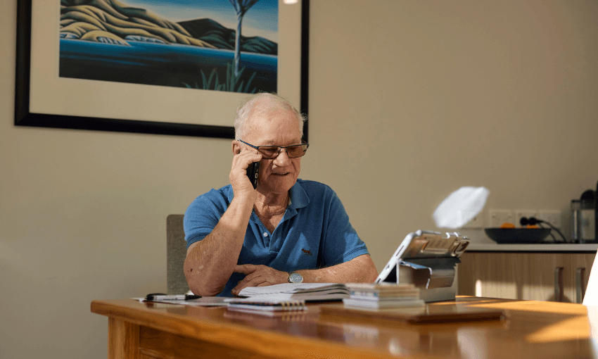 An older man wearing glasses and a blue polo shirt sits at a wooden table, talking on a smartphone. Papers, notebooks, and a tablet are in front of him. Sunlight streams in, and a landscape painting hangs on the wall behind him.