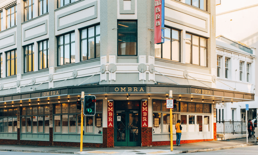 A person stands on a street corner outside Ombra, a restaurant in a light grey, three-story building with large windows and prominent signage, on a sunny day.