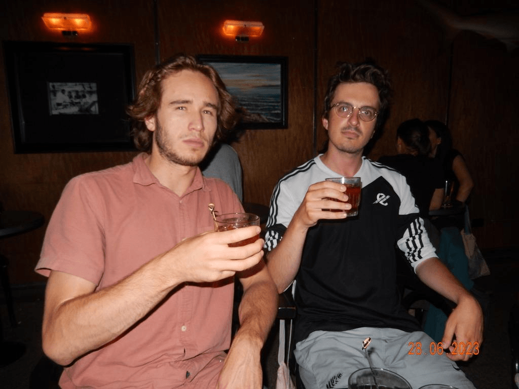 two young men holding cocktail glasses and sitting in a dimly lit bar