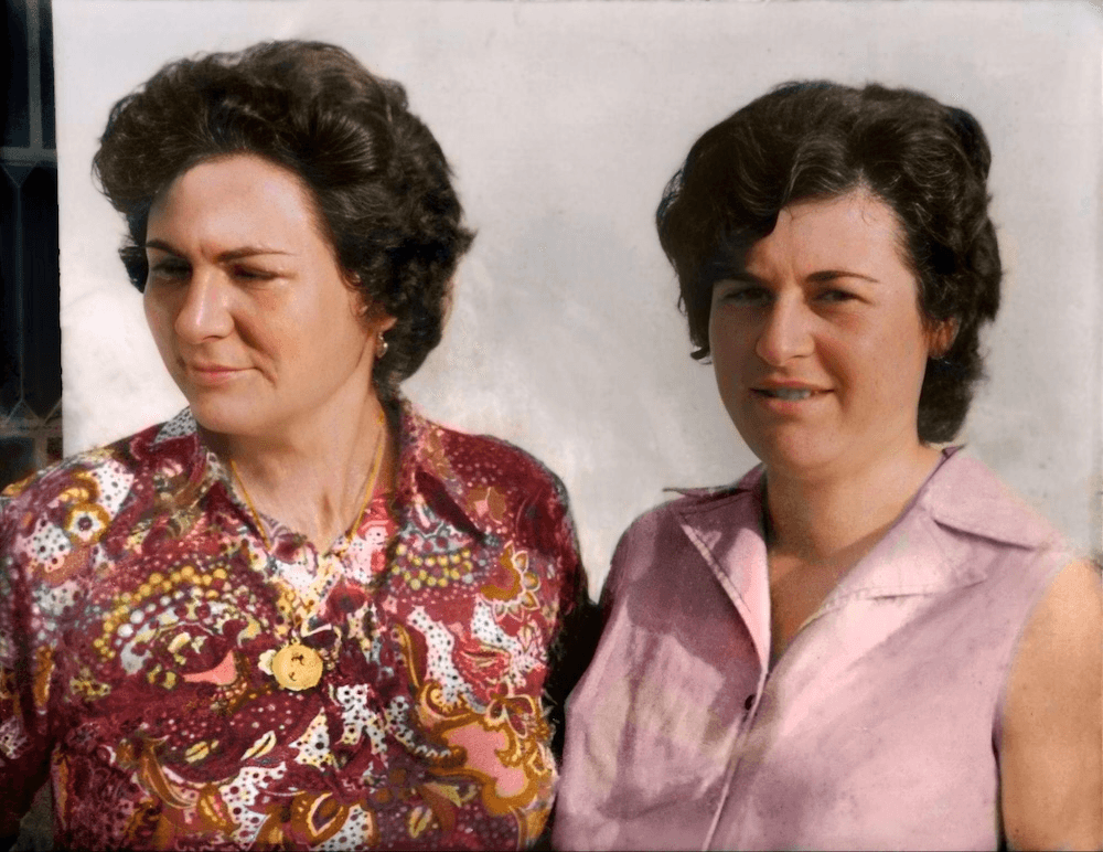 An old portrait of two Greek women with short dark, curly hair. one is frowning to the side and wearing a patterned blouse, the other is smiling to the camera and wearing a pink vest
