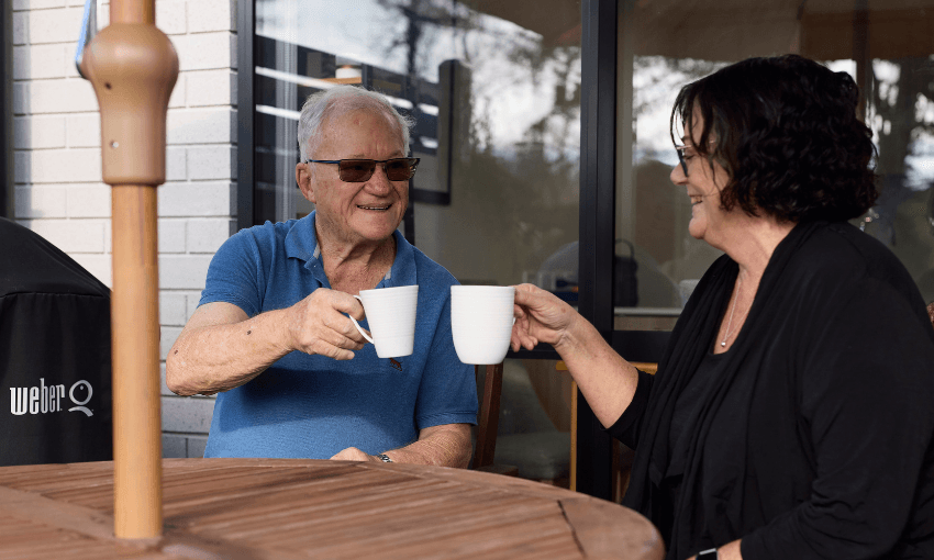 An older man and woman sit outside at a wooden table, smiling and clinking white mugs together. They appear to be enjoying a drink and conversation on a patio with a house and trees in the background.