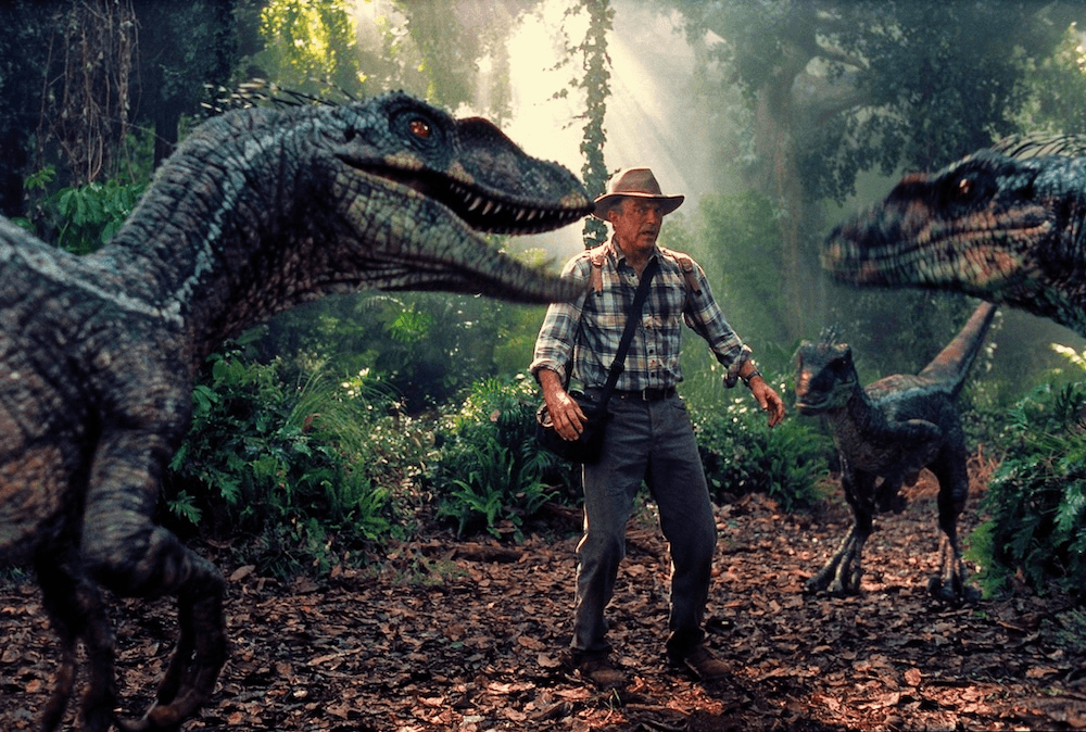 Actor Sam Neill in a scene from Jurassic Park III, standing in the middle of three dinosaurs in heavy greenery