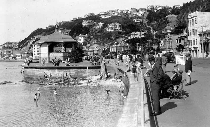 Black and white old photo of footpath and pedestrians looking down on people swimming in the water. There are houses in the hills in the distance