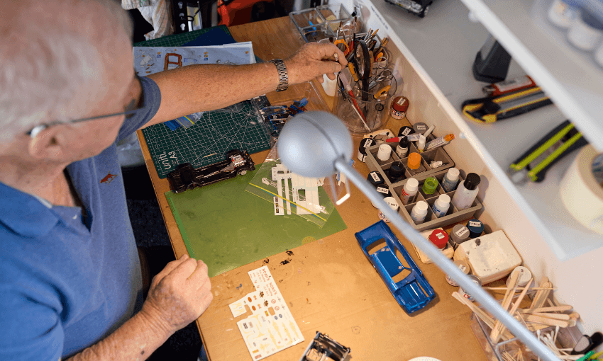 An older man works on assembling a model car at a tidy desk, surrounded by paints, tools, decals, and model car parts under a bright desk lamp.