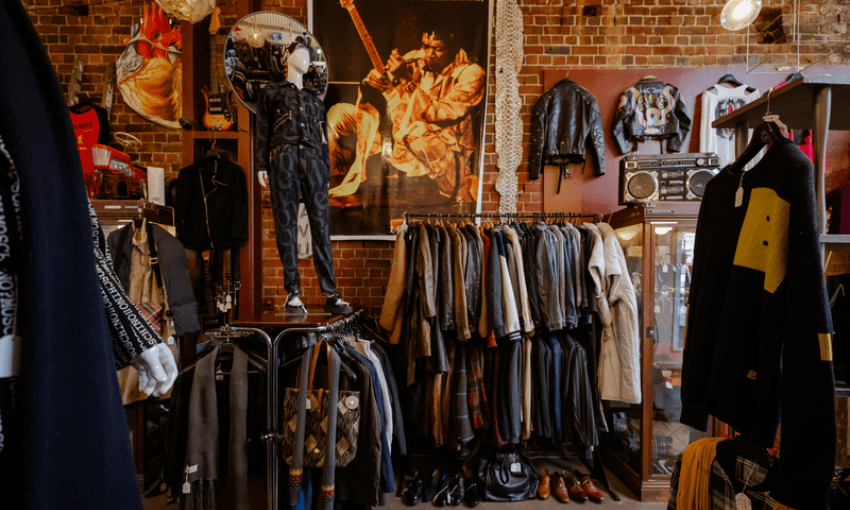 A vintage clothing store interior with racks of assorted jackets and shirts, a mannequin wearing a dark outfit, framed photos on brick walls, and a leather jacket and boombox displayed on shelves.