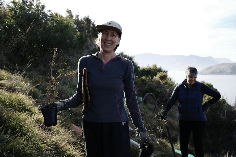 a woman with soalt an pepper hair smiling with a seedling in her hands and the sky behind her