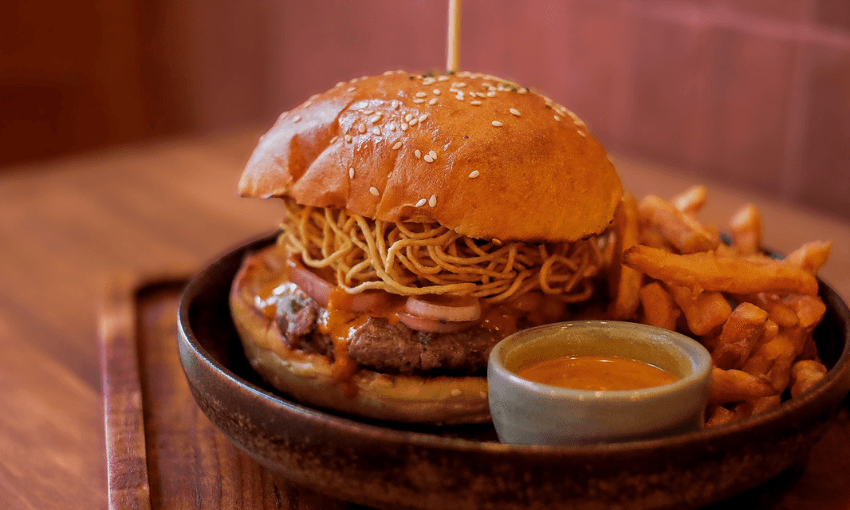 A close-up of a burger with crispy noodles, onions, and sauce in a sesame bun, served with French fries and a small bowl of dipping sauce on a wooden plate.