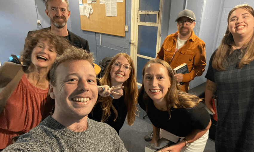 A group of seven smiling adults pose for a selfie in an indoor room with blue walls and a bulletin board in the background. Some people hold books, and one person flashes a peace sign.