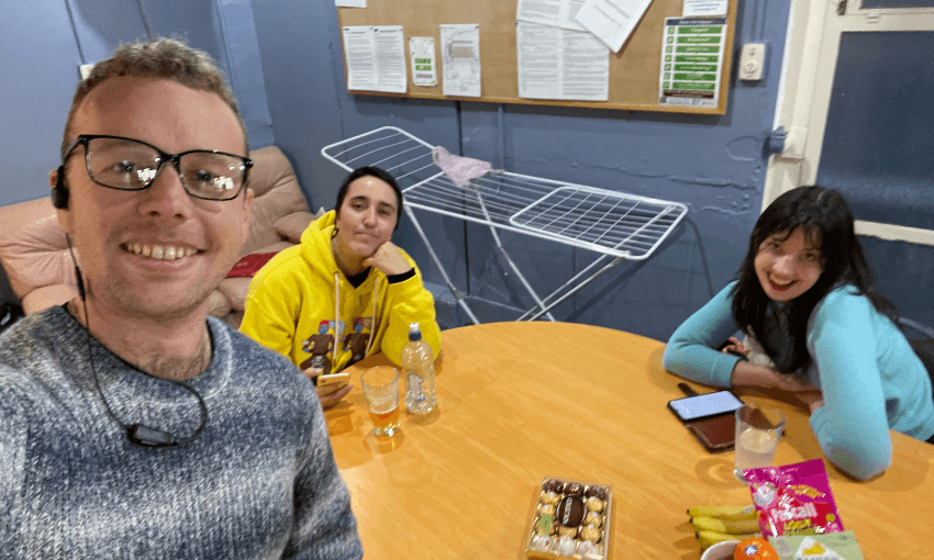Three people sit around a wooden table with snacks, drinks, and a phone. One person takes a selfie, smiling, while the other two also smile at the camera. An empty drying rack and bulletin board are in the background.