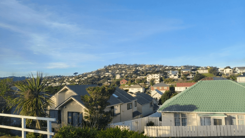 A sunny hillside neighborhood with many houses scattered across green slopes under a clear blue sky, with a few trees and fences in the foreground.